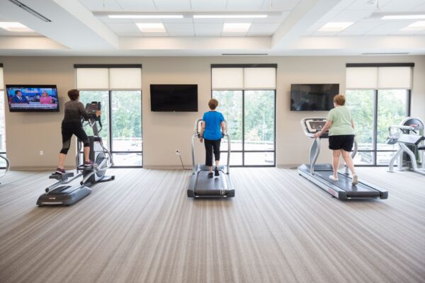 Three senior women exercising on treadmills in a bright and airy senior living gym.