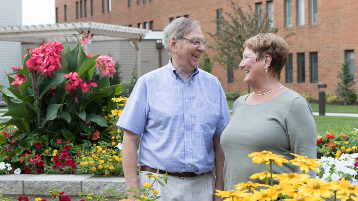 A senior man and senior woman gaze lovingly into each other's eyes while standing in a flower garden with bright yellow and pink flowers.